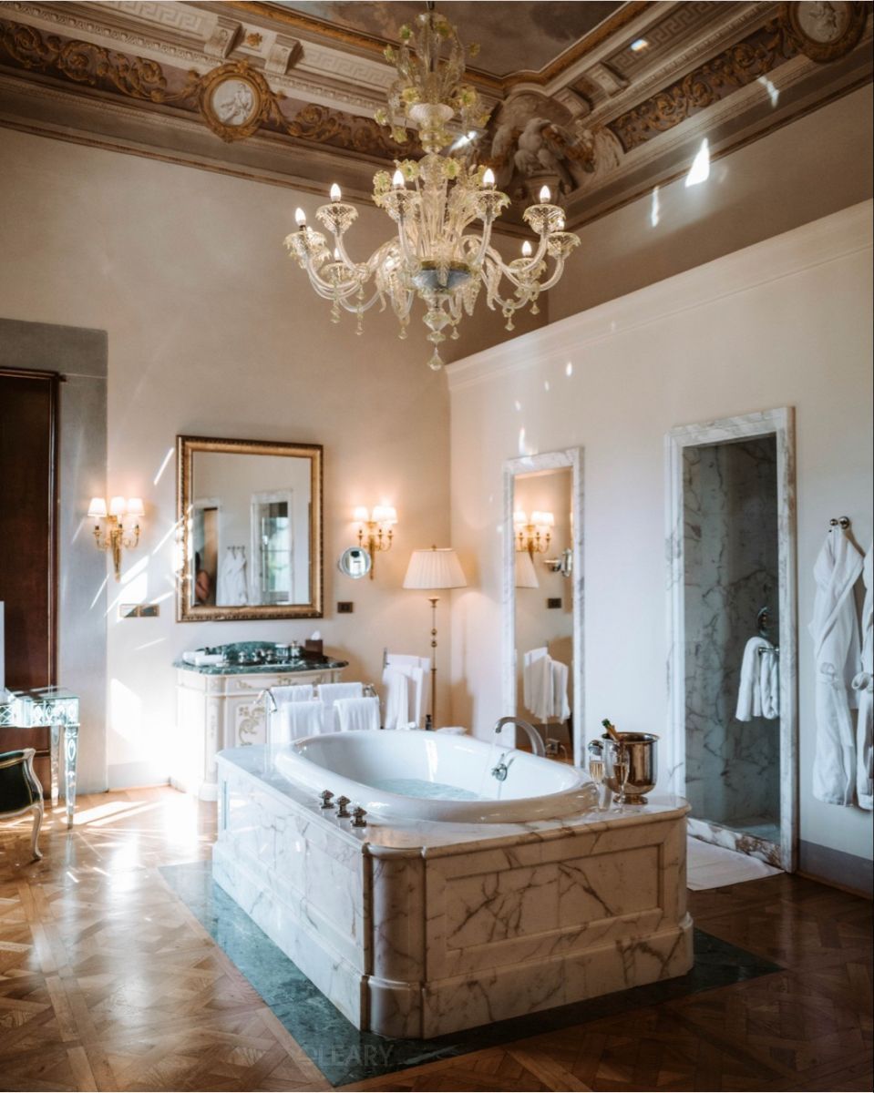 In a luxurious classical bathroom, a large Murano glass chandelier hangs from an ornate ceiling above a white marble built-in bathtub, with parquet flooring, creating a palatial and magnificent ambiance.