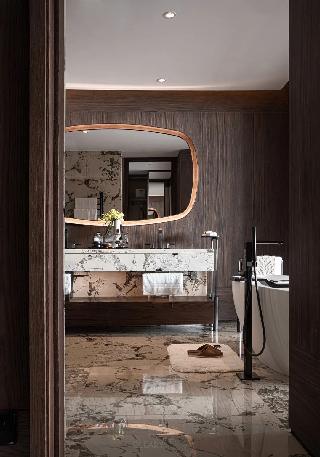 Opulent hotel bathroom featuring a stunning vanity with a thick, heavily veined marble washbasin and countertop on a dark wood and chrome frame, with a partial view of a pure white freestanding bathtub, all set against rich dark wood walls and dramatic marble flooring.