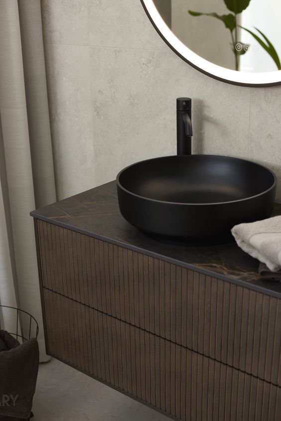 Sophisticated modern bathroom featuring a striking matte black round vessel sink on a dark stone-look countertop, mounted on a dark wood, vertically fluted floating bathroom cabinet, with a large backlit round mirror and a matching matte black faucet.