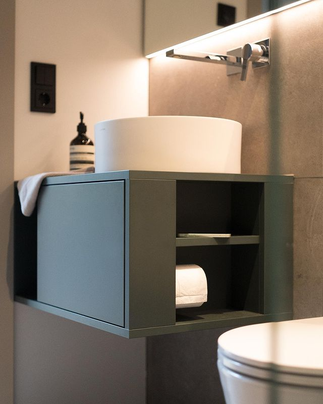 A modern bathroom featuring a white round solid surface countertop basin on a grey-green wall-mounted cabinet with open and closed storage, highlighting contemporary and functional design.