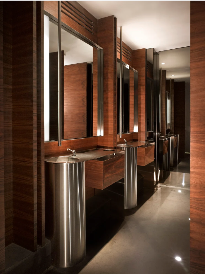A row of modern commercial pedestal sinks with brushed stainless steel bases and dark wood vanities in a luxury hotel restroom, showcasing sleek design and durability.