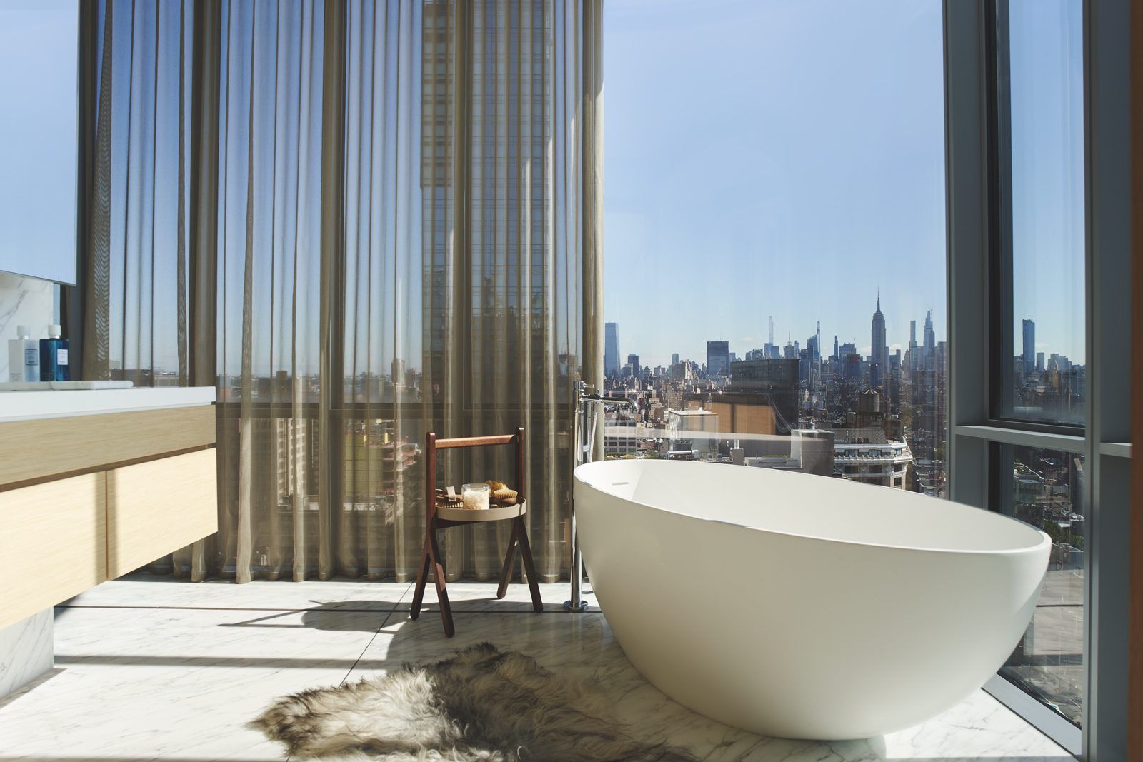 A luxurious modern bathroom featuring a white freestanding oval bathtub with a city view, a wooden side table stool holding a tray, and a plush fur rug on a marble floor, creating a serene spa-like atmosphere.