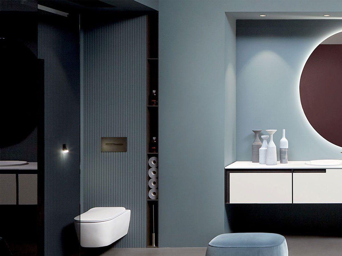 A sophisticated modern bathroom with blue-grey walls, a wall-mounted toilet in a fluted panel alcove, and a sleek floating vanity with a white countertop/basin and a large backlit round mirror.