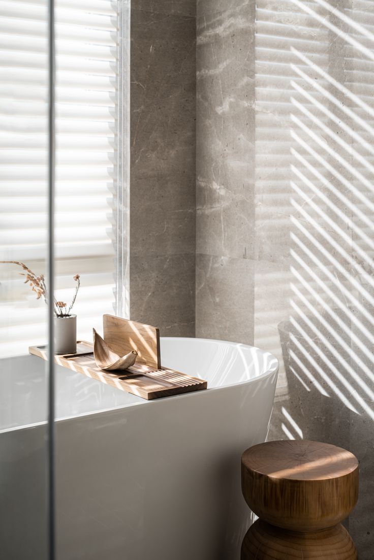 A serene spa-like bathroom with a white freestanding bathtub, a wooden bath caddy holding botanicals, a round wooden stool, and dramatic light stripes from window blinds on stone-look tiled walls.
