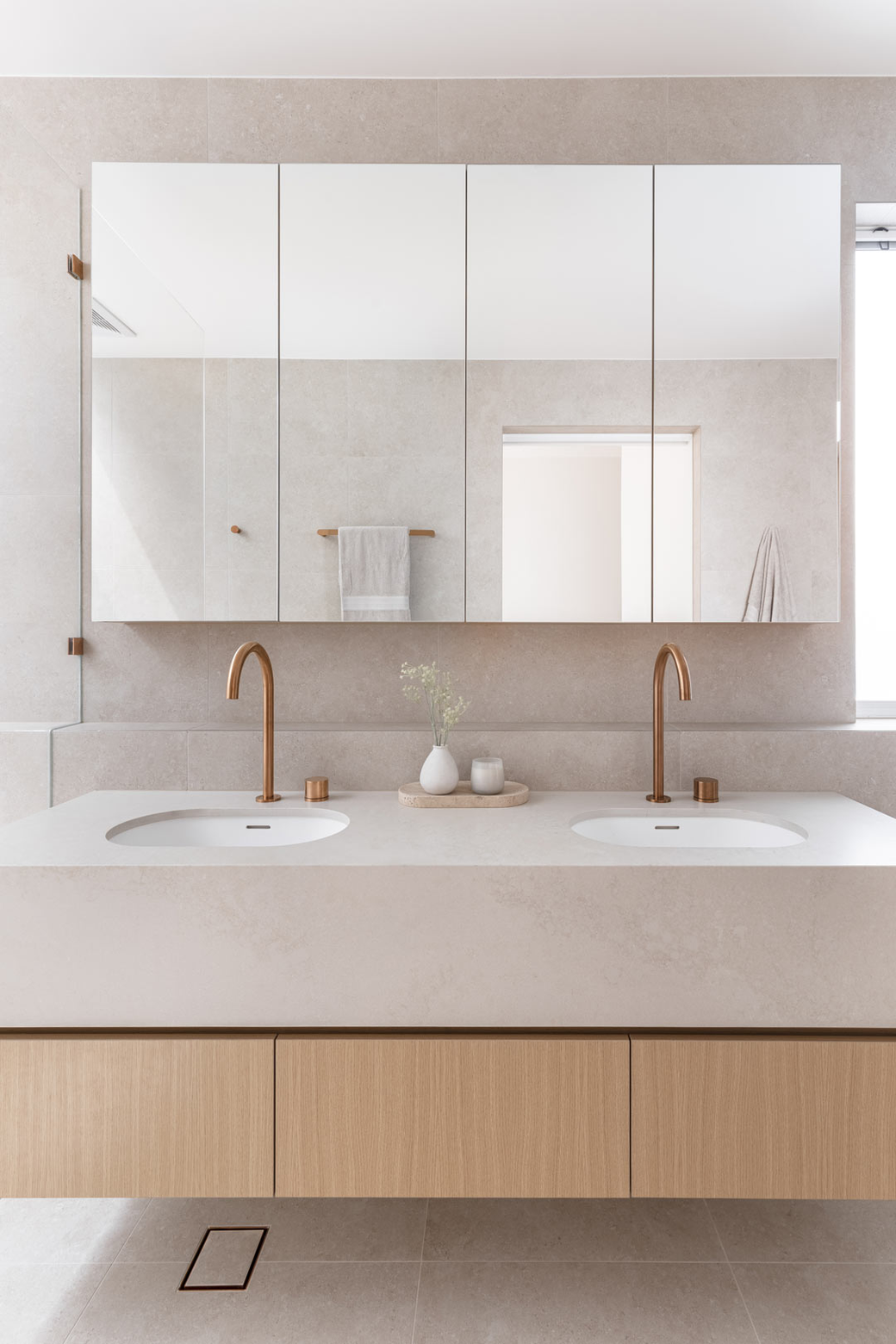 A serene modern bathroom featuring a light wood floating double vanity with a white countertop and undermount sinks, topped by a large multi-panel mirror cabinet, with copper faucets and light tiled walls.