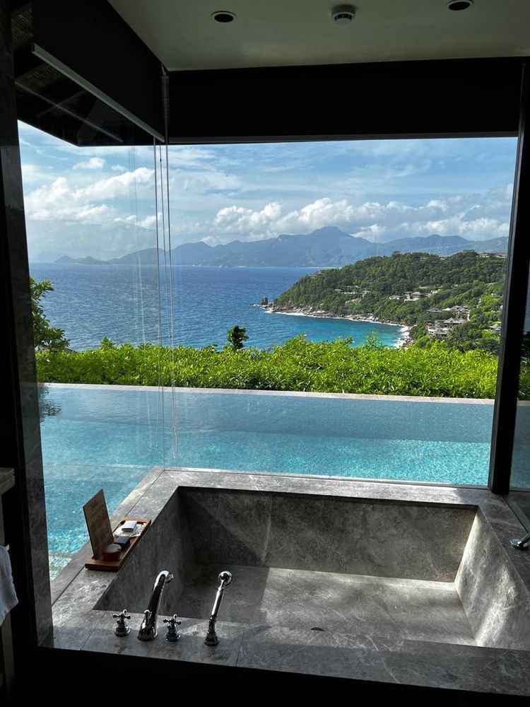 Luxurious hotel bathroom featuring a dark marble embedded bathtub overlooking a private infinity pool and a breathtaking tropical ocean and mountain vista, epitomizing a dream resort vacation.