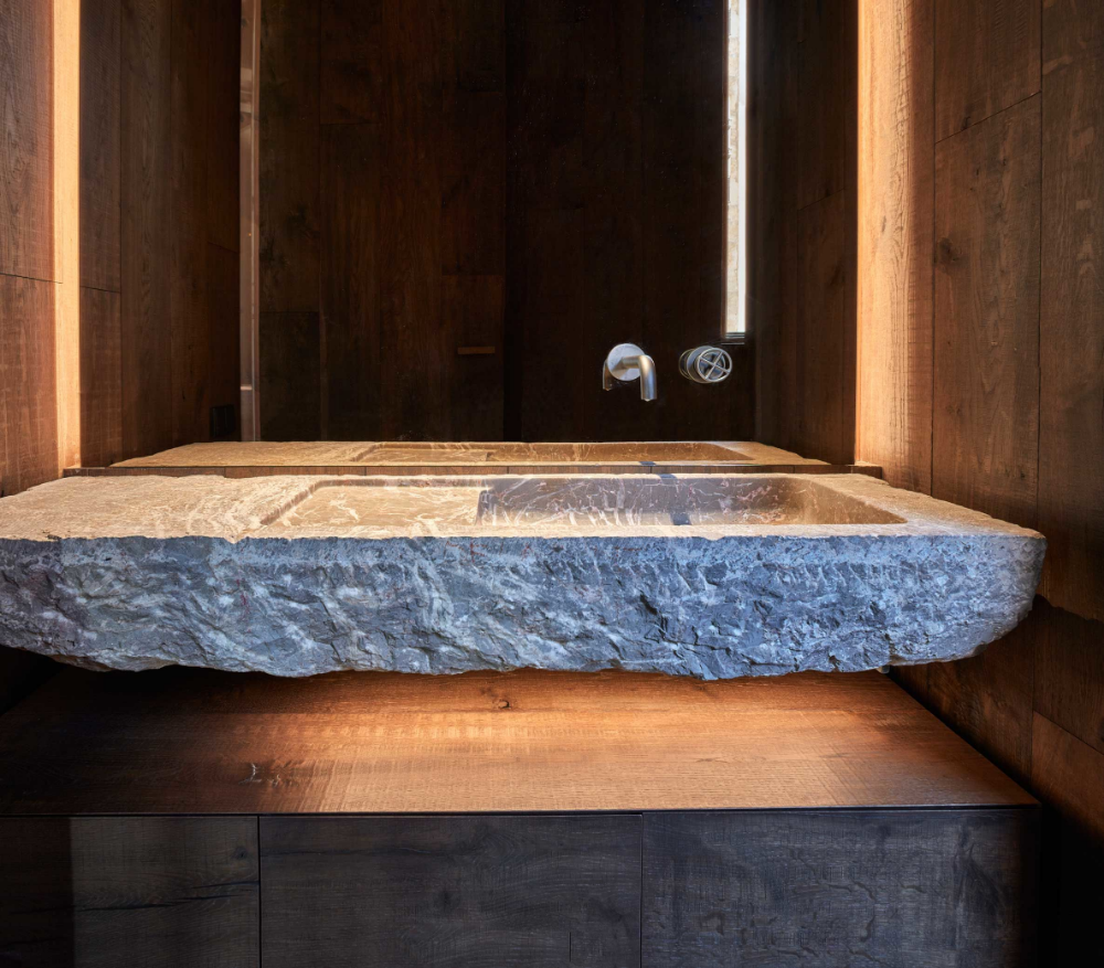 Dramatic rustic-luxe bathroom featuring a massive grey natural stone trough sink with a rough-hewn front, mounted on a dark wood vanity, with dark wood paneled walls and atmospheric strip lighting.