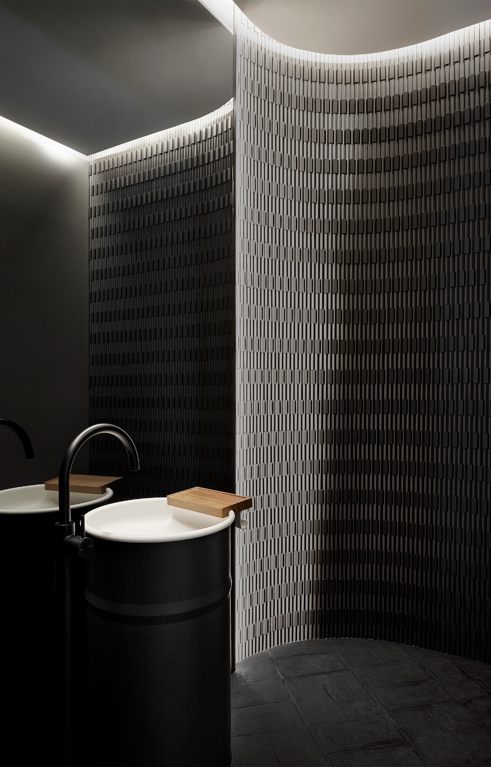 Two modern black industrial-style pedestal sinks (Agape Vieques style) with white basins and matte black gooseneck faucets, set against a dramatically lit, curved, textured tile wall in a luxury commercial bathroom.