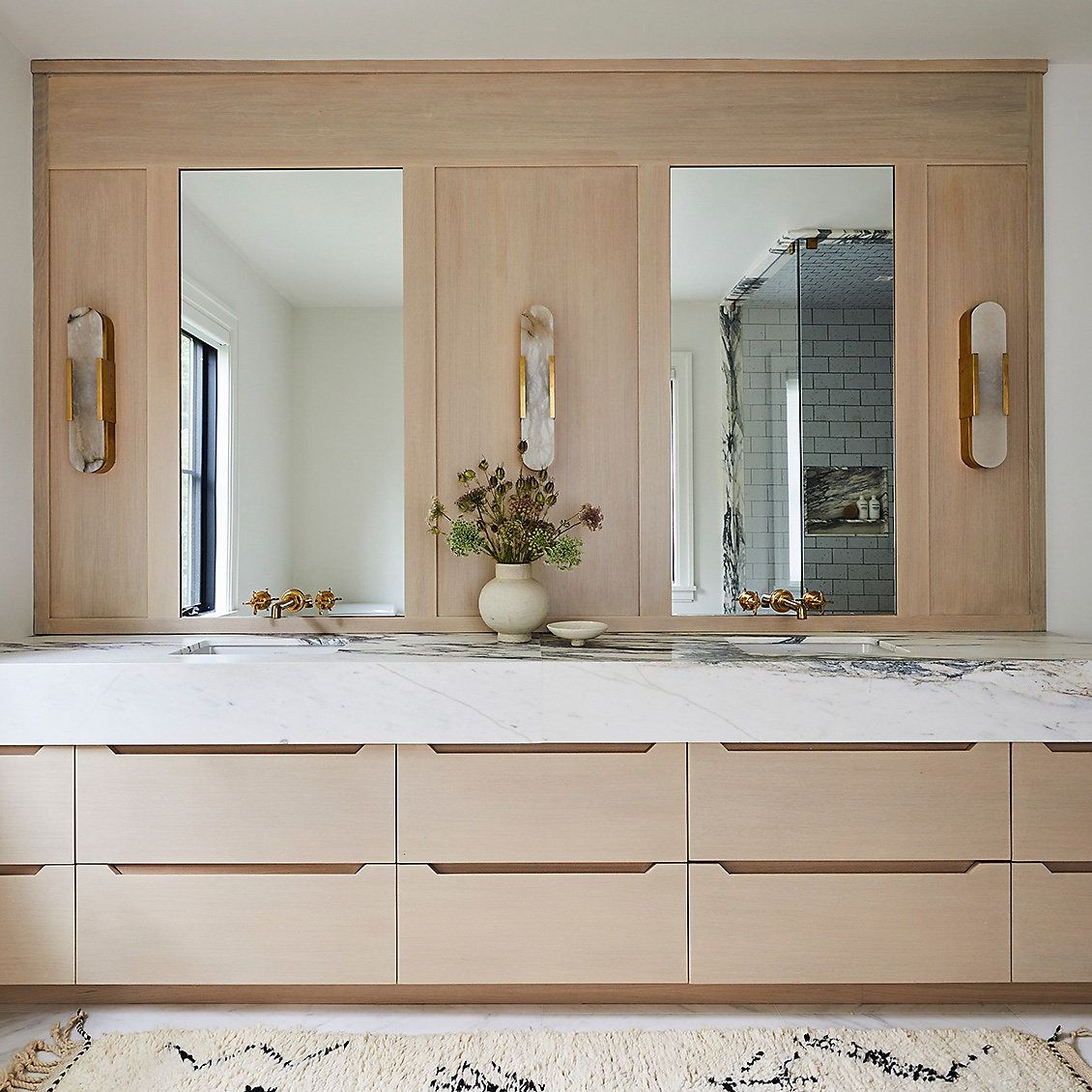 Sophisticated hotel-inspired bathroom featuring a large, light wood floor-standing double vanity with a pristine white marble countertop, elegant gold faucets, and ambient wall sconce lighting.