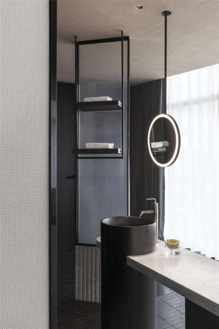 Chic modern bathroom featuring a bold black matte round seamless pedestal sink, complemented by a circular LED pendant light and reeded glass shelving in a sophisticated, moody setting.