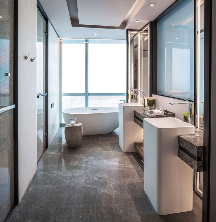Opulent hotel bathroom featuring a pristine white freestanding bathtub against a panoramic window, complemented by dual modern square pedestal washbasins with dark stone accents and vertically lit mirrors.