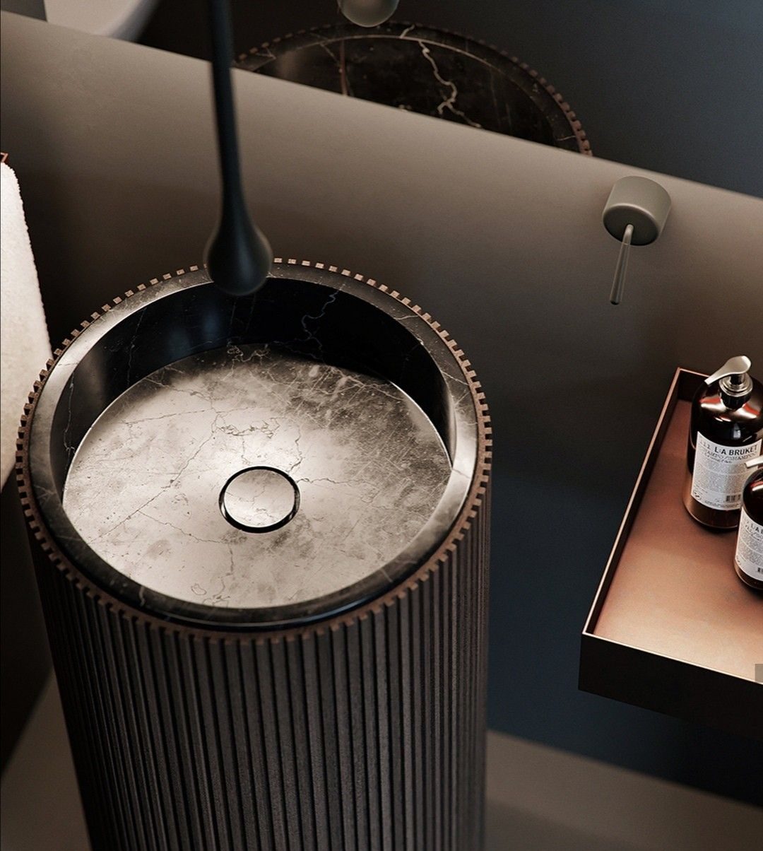 Exquisite close-up of a luxury tall pedestal sink, featuring a dark fluted cylindrical base with a decorative studded rim, an inset dark marble basin, and a striking matte black arched faucet, in a sophisticated, moody bathroom setting.