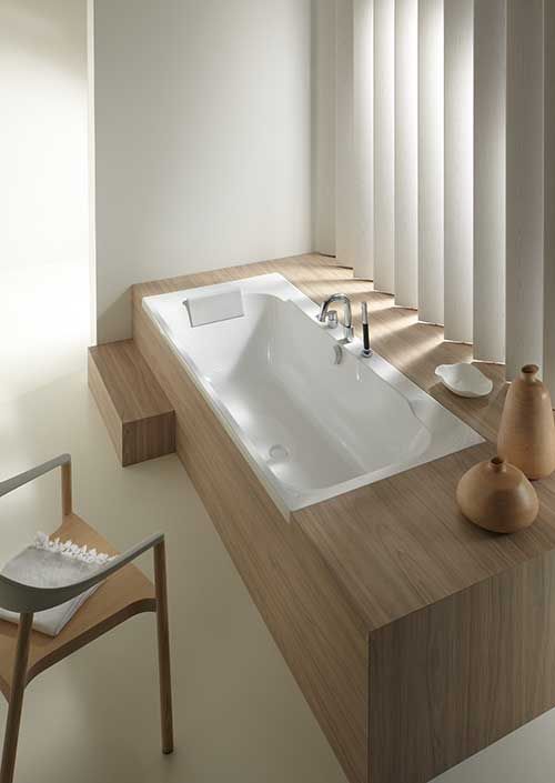 Tranquil modern bathroom featuring a pristine white glossy built-in bathtub with an expansive light wood surround, complemented by chrome deck-mounted faucets and soft, diffused light from vertical blinds, evoking a spa-like serenity.