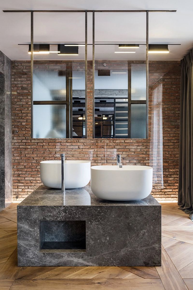 Striking industrial-chic bathroom featuring two white oval vessel sinks atop a dark grey marble island washstand, with tall chrome faucets, against an exposed red brick wall and illuminated by a suspended metal-framed mirror and lighting unit.
