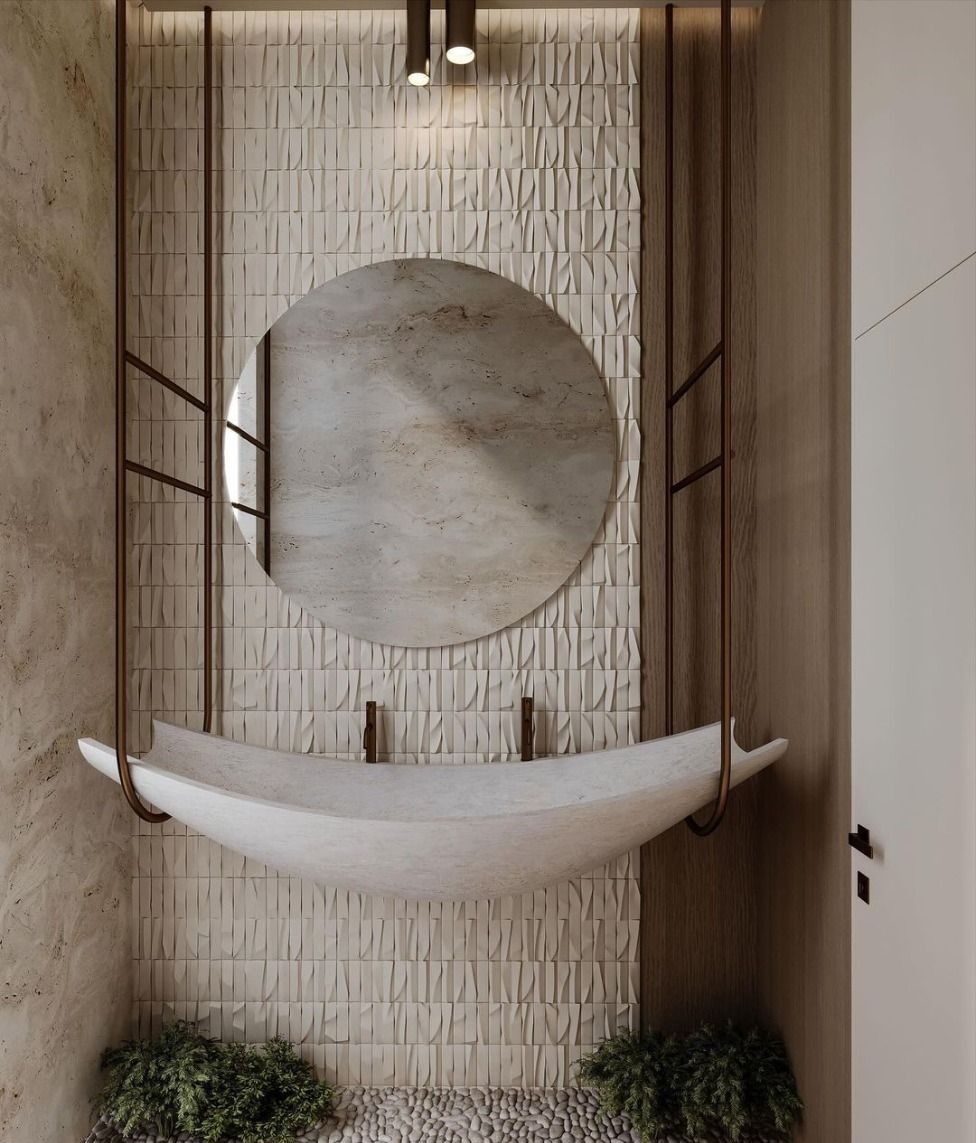 Artistic powder room featuring a stunning, boat-shaped, light stone custom washbasin suspended by a dark metal frame, with minimalist wall-mounted faucets, against a textured tile wall, a round textured mirror, and a bed of pebbles below.