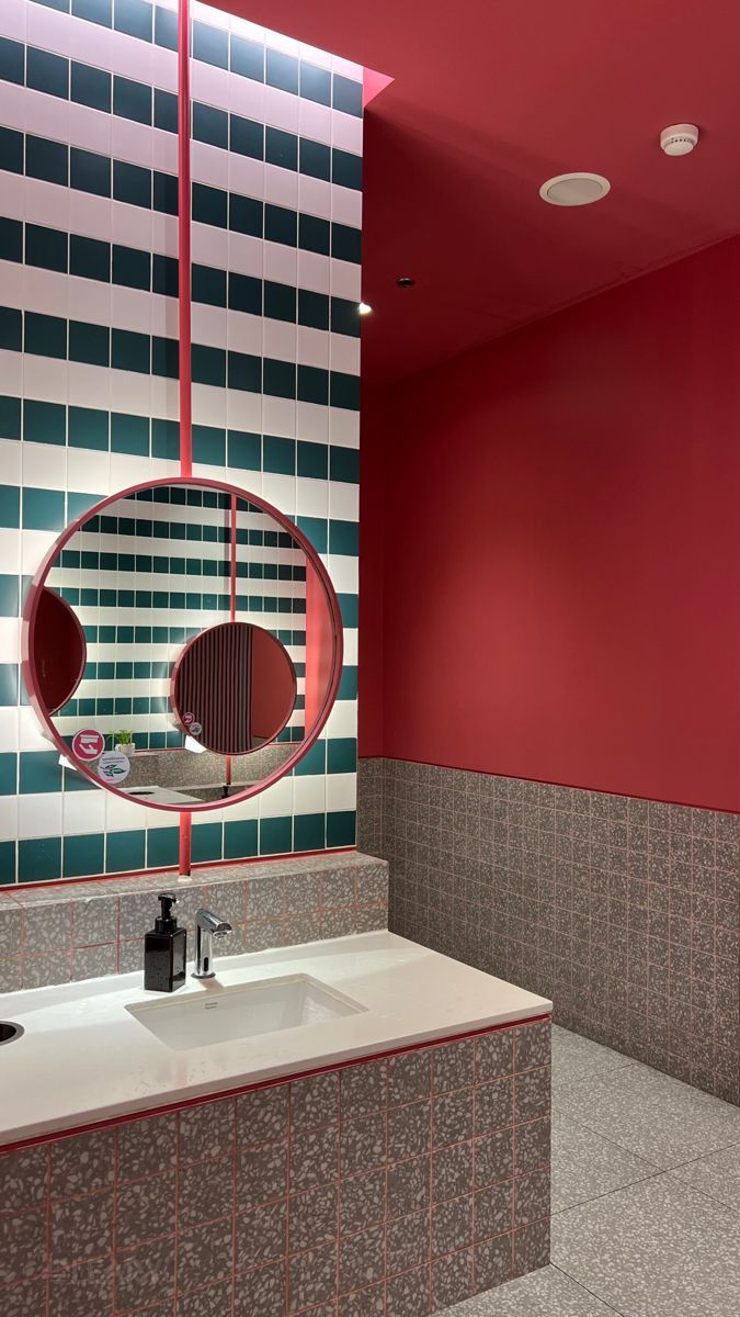 Modern bathroom wash area with a white undermount sink in a terrazzo-patterned vanity, against a teal and white striped tile wall, featuring a large round mirror with a bright red frame.