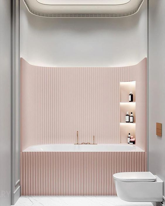 Pink built-in bathtub in a modern luxury bathroom, surrounded by vertical fluted paneling, with illuminated niches on the right wall and accented by gold faucets, creating a warm and tranquil ambiance.