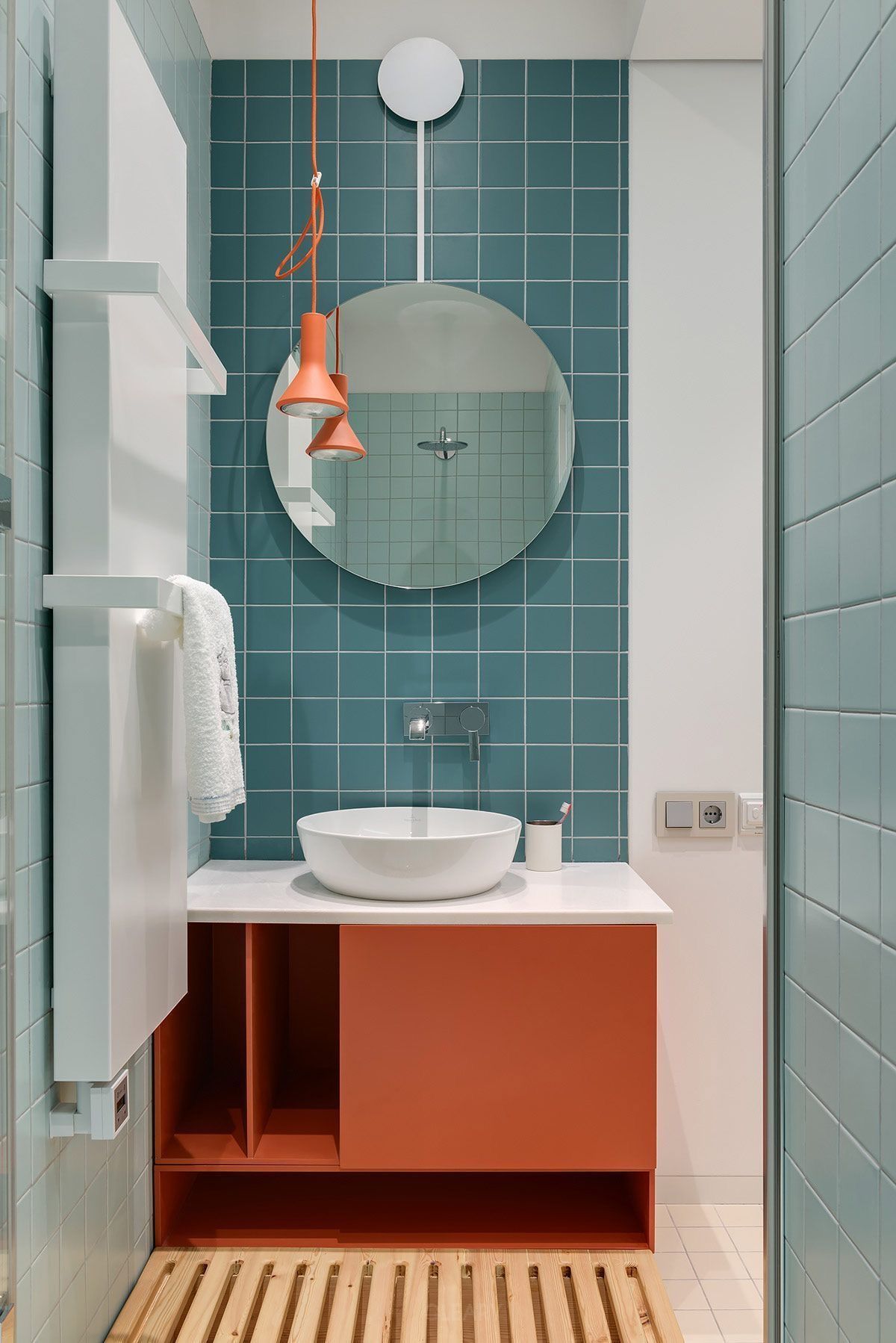 Modern bathroom featuring a white round vessel sink on a terracotta vanity, against a teal square-tiled wall, with a round mirror and an orange pendant light.