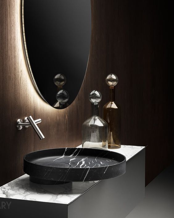 Close-up of a luxurious modern bathroom: a black marble round vessel sink on a white marble countertop, against a dark wood-paneled wall with a large oval backlit mirror, and a sleek wall-mounted faucet.