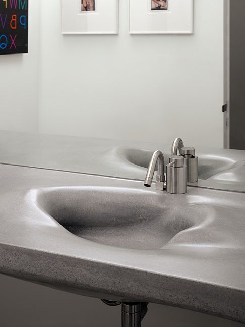 A grey concrete integrated sink with an irregular wave-shaped basin, fitted with a brushed stainless steel faucet, set against a white wall and mirror, showcasing modern industrial and artistic design.