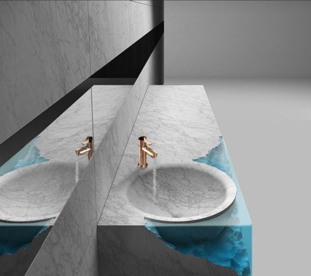 A modern artistic washbasin featuring a white marble circular basin and countertop, supported by an irregularly shaped transparent blue resin base, with a rose gold faucet, against a marble wall and mirror.