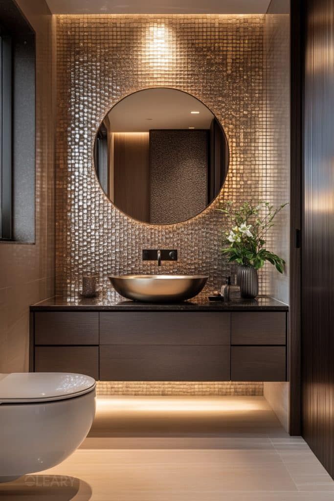 A luxurious modern bathroom featuring a bronze metallic countertop basin on a dark stone countertop with a wood grain vanity, set against a gold mosaic tile accent wall, complete with a round mirror and ambient lighting.