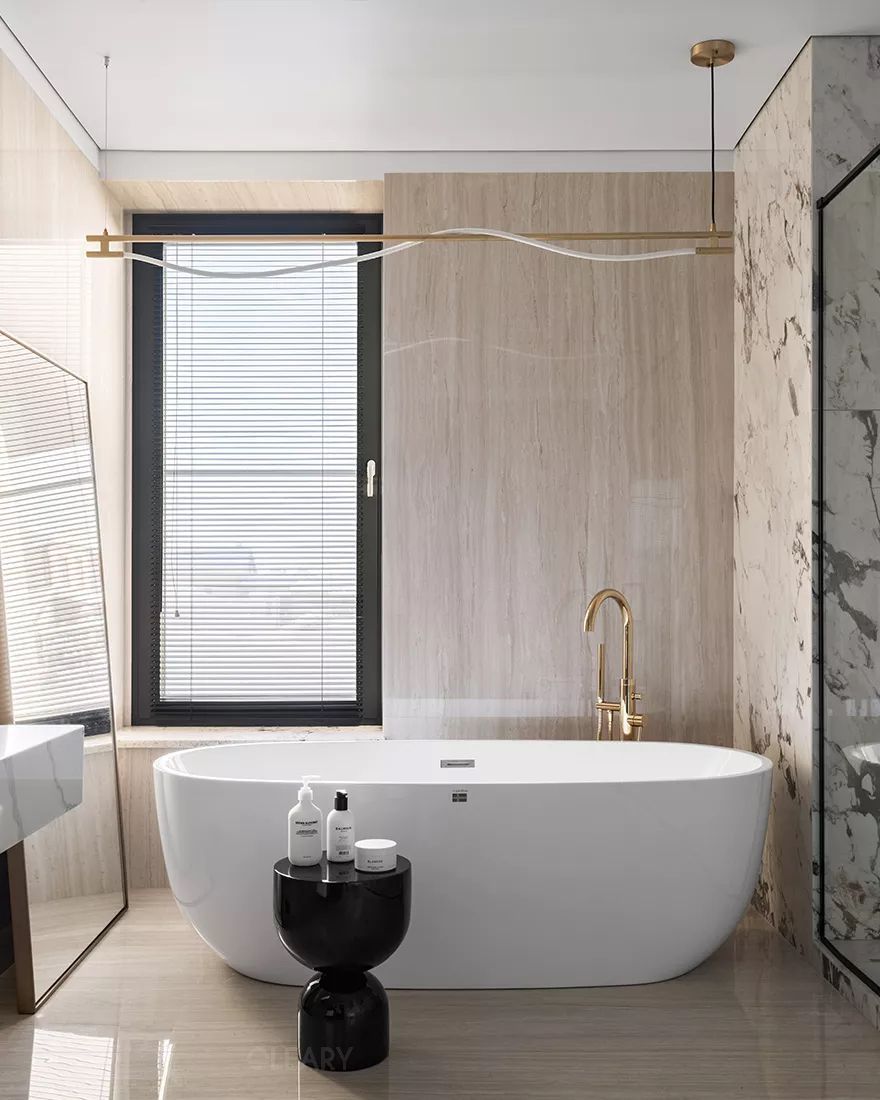 A modern luxury bathroom features a sleek white freestanding acrylic bathtub by a bright window, complemented by a gold floor-mounted faucet and a black side table, showcasing style and comfort.