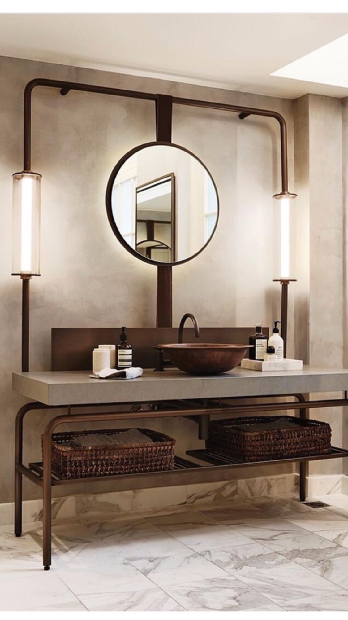 A modern industrial-style bathroom featuring a dark metal frame vanity with a concrete countertop, a bronze round vessel sink, and a round mirror with LED backlight and flanking pipe-style wall sconces.