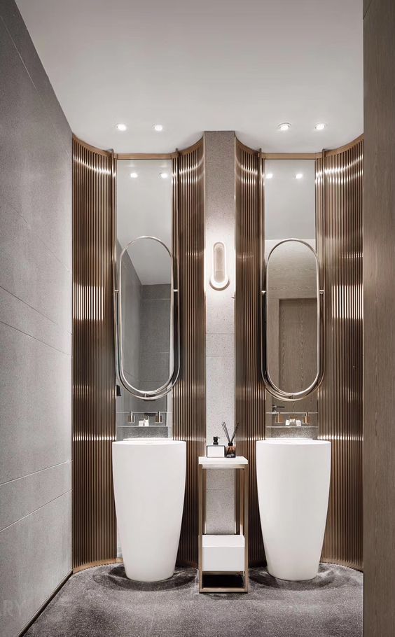 A modern luxury double vanity area with two white round freestanding pedestal sinks, set against a wavy bronze ribbed panel wall, with oval metallic-framed mirrors and a wall sconce above.