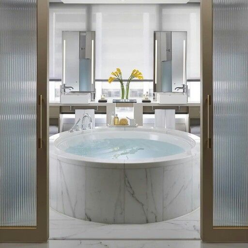 A luxurious marble bathroom featuring a large central round bathtub, with a double vanity unit with large illuminated mirrors in the background. (Note: This alt text describes the main elements in the image but does not emphasize 