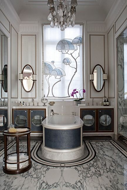 Classical luxury hotel bathroom featuring a central oval bathtub with artistic cladding, flanked by custom dark wood vanities, with marble flooring and inlay patterns.

