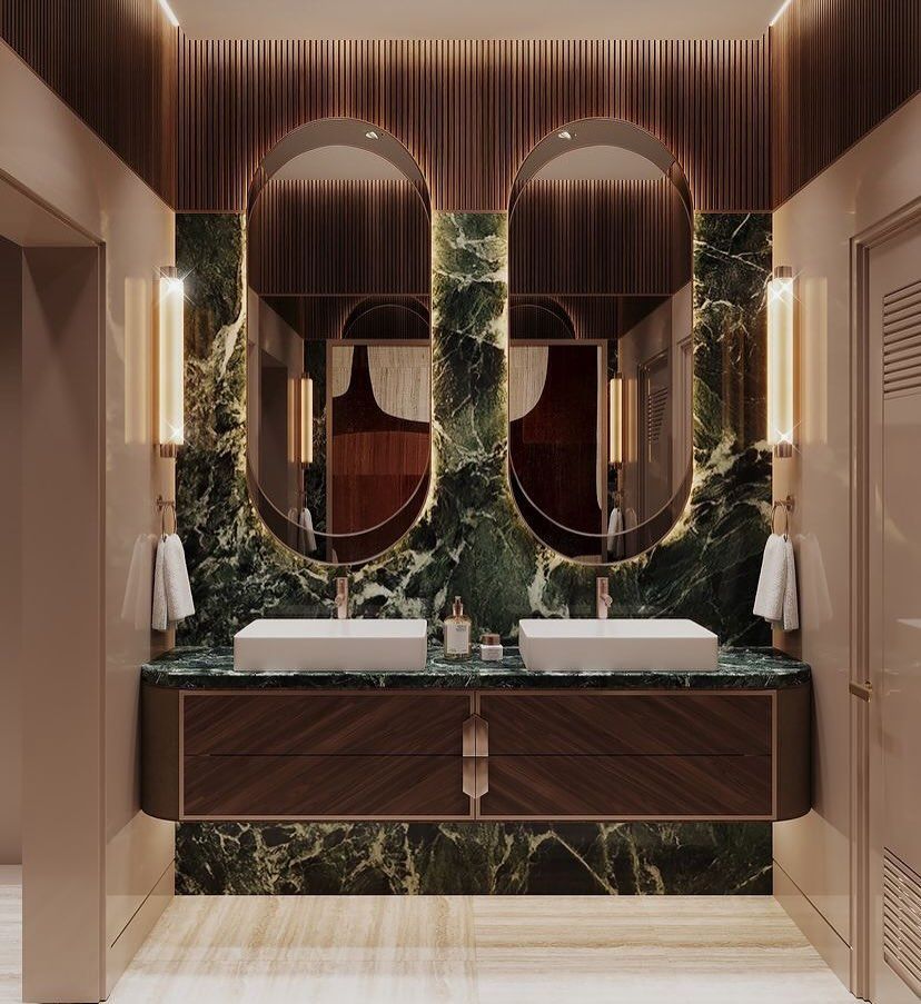 A luxurious Studia 54 bathroom with a dark wood floating vanity, two white rectangular vessel sinks on a green marble backsplash, and two large oval brass mirrors flanked by glowing sconces.