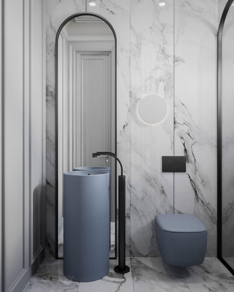 A modern bathroom featuring a matte blue-gray round pedestal sink with a tall black floor-mounted faucet, set against a white marble wall with a large arched black-framed mirror.
