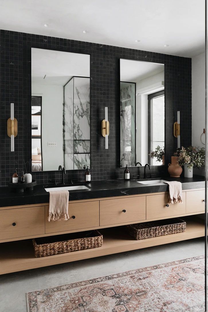 A modern floating wood double vanity with a black countertop and white undermount sinks, set against a matte black square tile wall with vertical sconces.