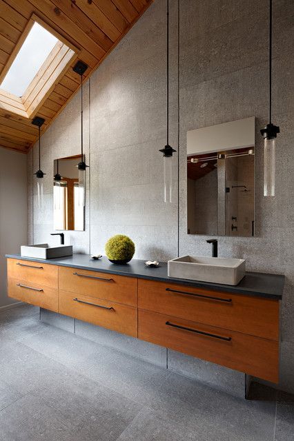 A modern spa-like bathroom with a long, floating wood double vanity, grey concrete vessel sinks, and pendant lights hanging from a vaulted, wood-paneled ceiling with a skylight.