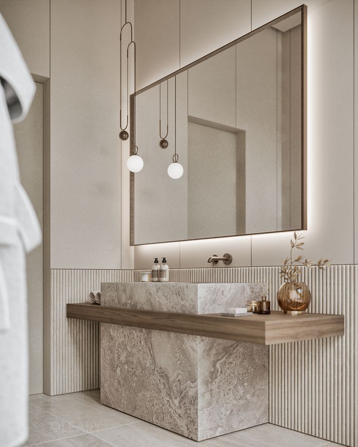 A serene, minimalist bathroom featuring a monolithic beige marble vanity with an integrated sink, a floating wood shelf, a large backlit mirror, and designer pendant lights against a fluted wall.