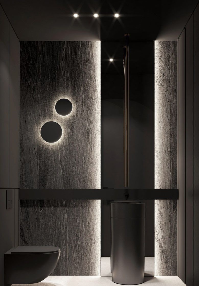A moody, minimalist bathroom with a dark grey freestanding pedestal sink, highlighted by vertical backlighting on textured stone walls and circular wall sconces.