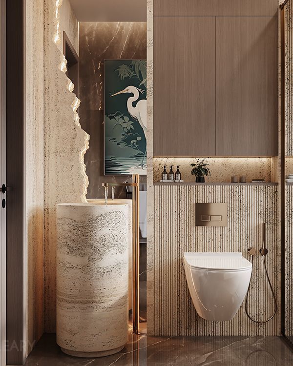 A luxurious bathroom featuring a cylindrical travertine stone pedestal sink with a floor-standing brass faucet, a wall-hung toilet, and warm, layered lighting that creates a sophisticated and spa-like ambiance.