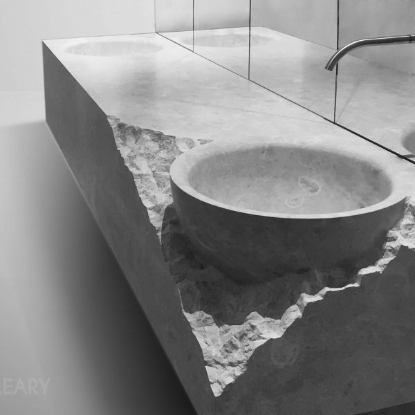 A monolithic stone washbasin featuring a smooth, integrated round bowl that contrasts with a raw, chiseled front edge, showcasing brutalist-inspired bathroom design in black and white.