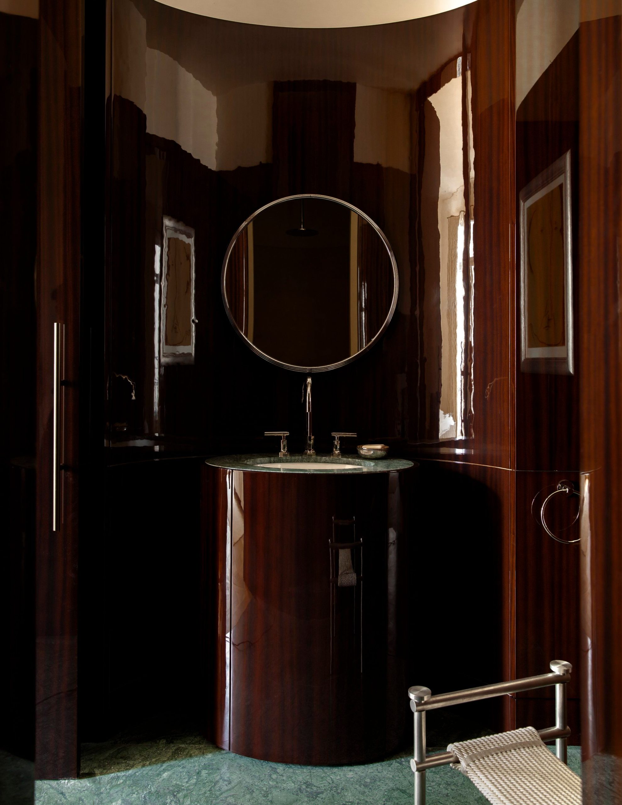 An ultra-luxurious Art Deco bathroom featuring a cylindrical high-gloss wood pedestal sink with a green marble top, chrome fixtures, and enveloping curved wood walls.