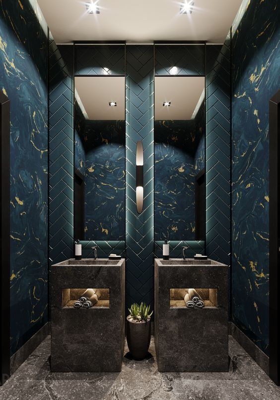 A symmetrical luxury powder room featuring two rectangular dark stone pedestal sinks, a teal herringbone tile accent wall, and dramatic teal and gold marble walls.