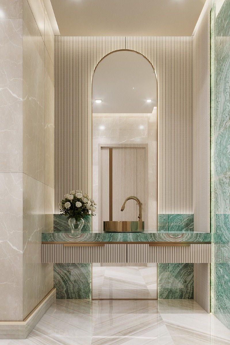 A luxury bathroom featuring a round brushed brass vessel sink on a green marble floating vanity, with a tall arched mirror and a cream-colored fluted wall behind it.