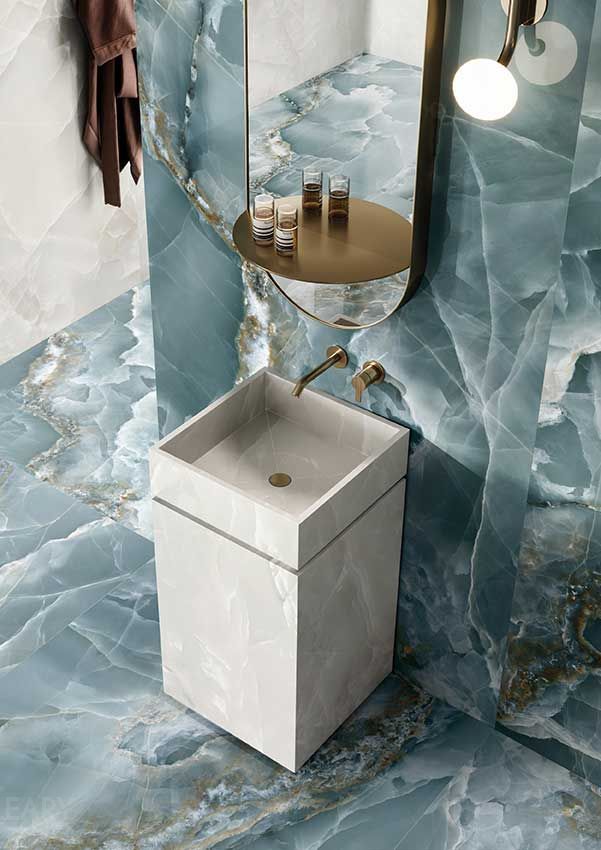 A modern, white, rectangular pedestal sink stands against a dramatic wall of blue and gold onyx-effect tiles, complemented by a wall-mounted brass faucet and an oval mirror.