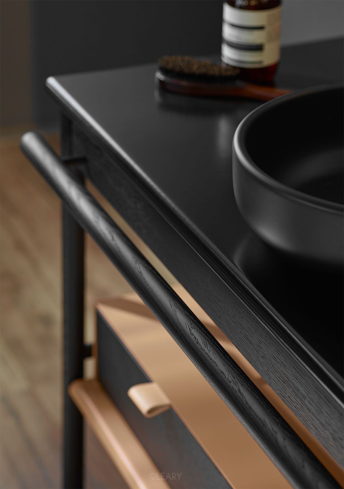 A close-up of a sophisticated matte black custom vessel sink resting on a dark solid oak bathroom vanity, detailed with a natural wood drawer and leather pull.