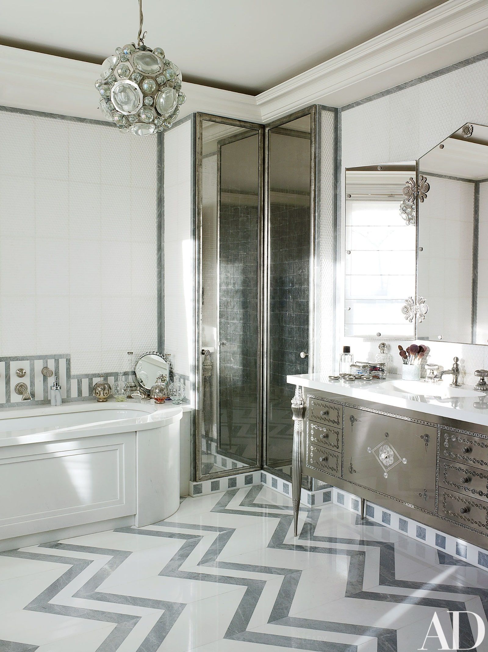 A glamorous bathroom designed by Jacques Grange, featuring a silver metallic furniture-style vanity with elegant legs on a bold grey and white chevron marble floor.