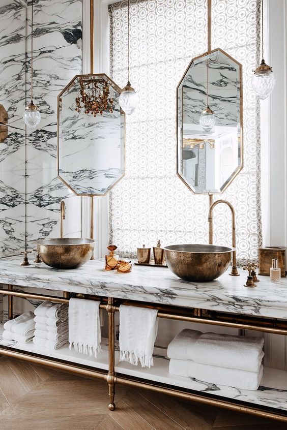 An elegant bathroom with a long marble-topped washstand, featuring two bronze vessel sinks, tall brass faucets, and octagonal mirrors against a book-matched marble wall.