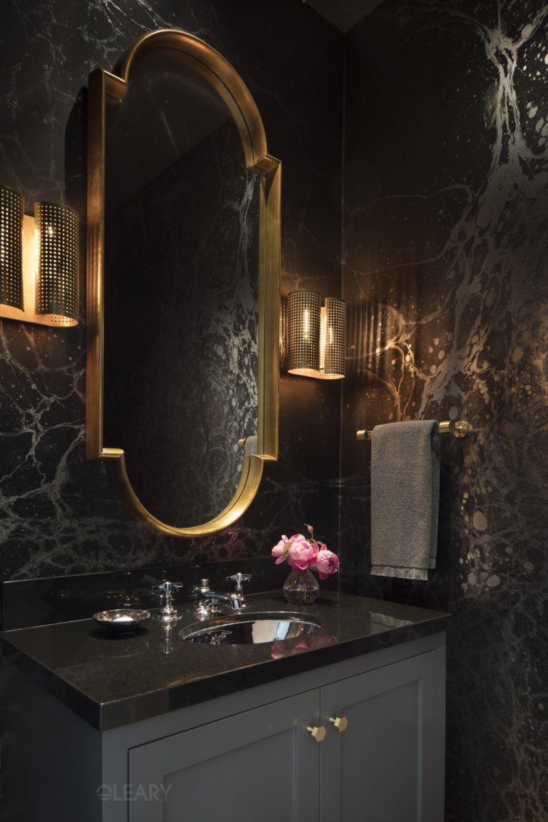 A luxurious powder room featuring a charcoal grey bathroom vanity with a black stone countertop, an ornate gold-framed mirror, elegant gold wall sconces, and dramatic black marble walls with gold veining.