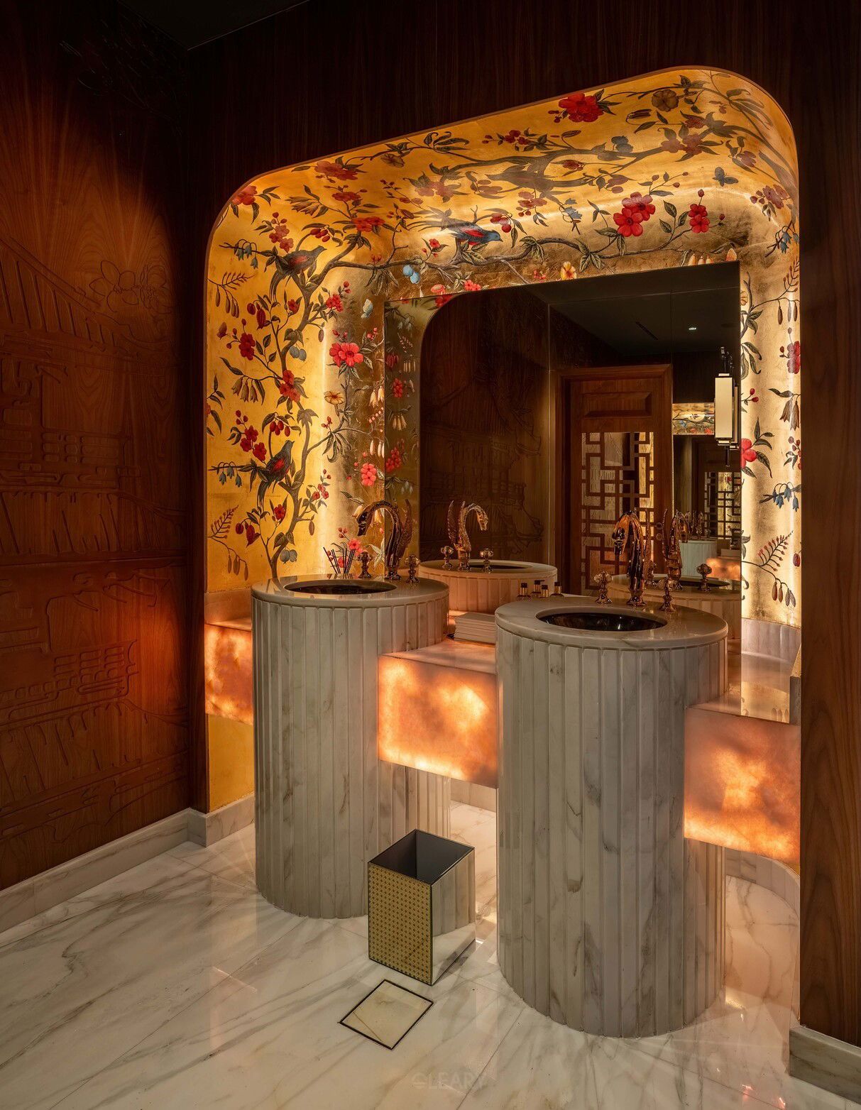Opulent Chinoiserie-themed luxury bathroom featuring two fluted marble pedestal sinks with dark contrasting basins, set against backlit onyx panels, within a grand archway lined with gold-leaf floral and bird motif wallpaper.