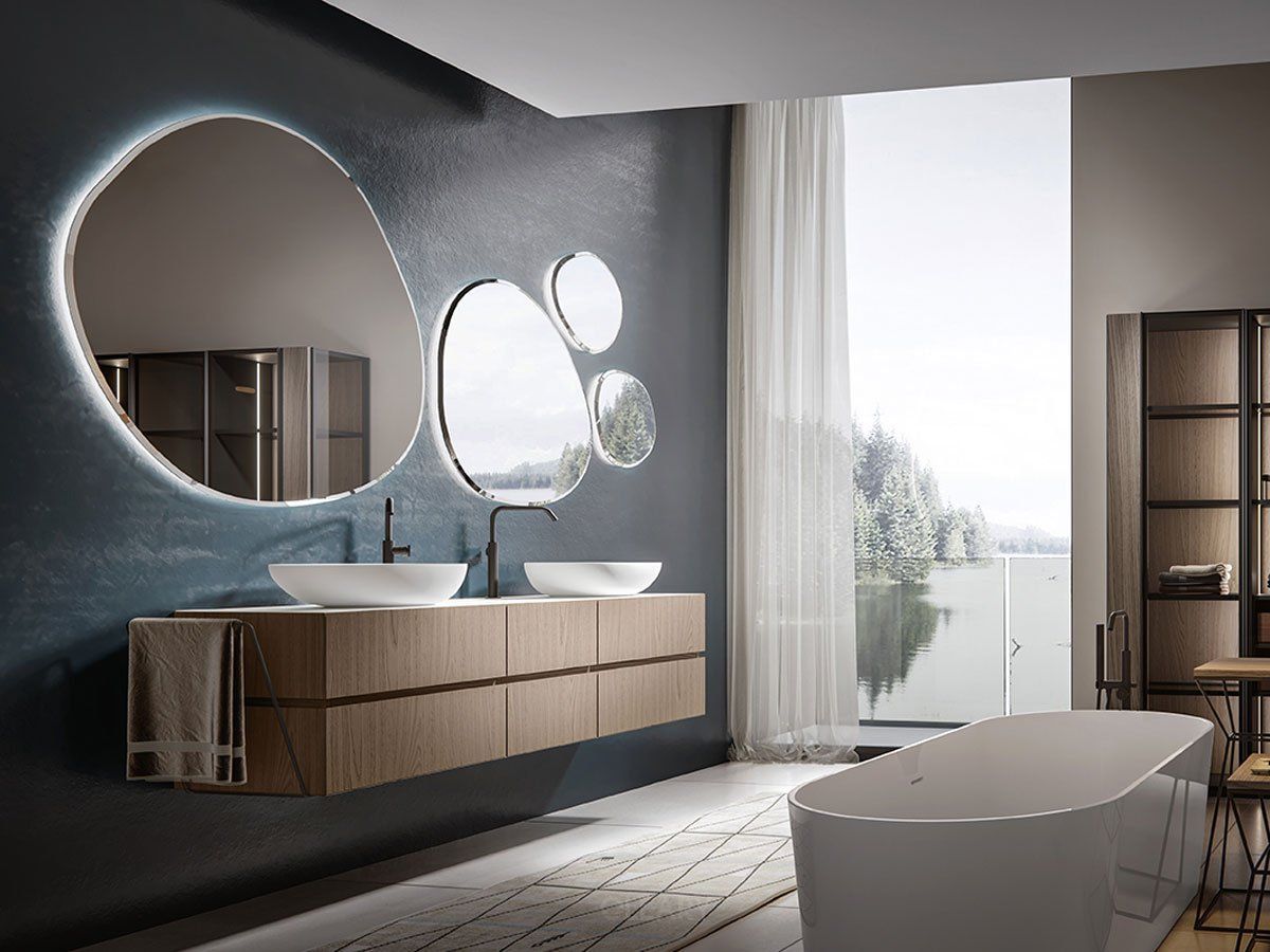 Serene modern luxury bathroom featuring a long floating wood vanity with two white vessel sinks, matte black faucets, a set of organic-shaped backlit mirrors, a white freestanding bathtub, and a large picture window with a tranquil forest and lake view.