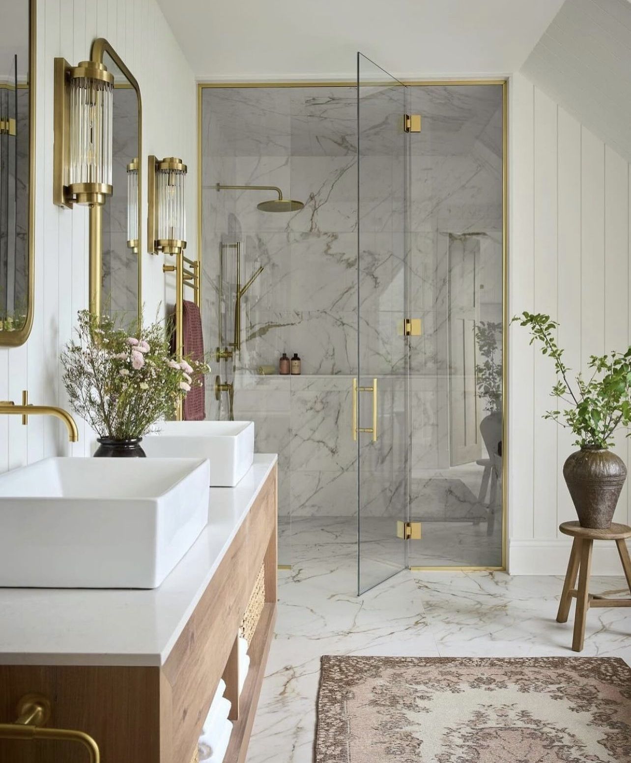Elegant transitional bathroom featuring a light wood double vanity with two white rectangular vessel sinks, wall-mounted gold faucets, twin gold-framed mirrors with rounded tops, and a luxurious marble-tiled shower with gold hardware.